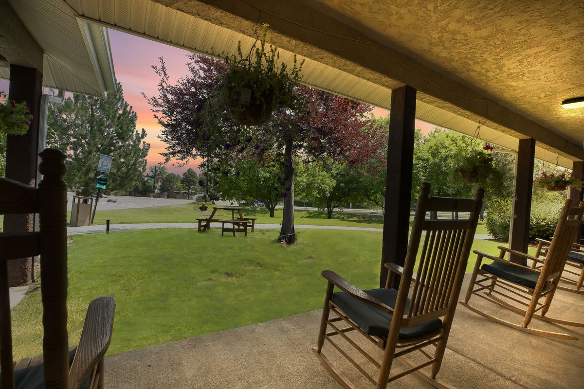 A tranquil evening view from a porch with rocking chairs, overlooking a lush lawn and trees at sunset.