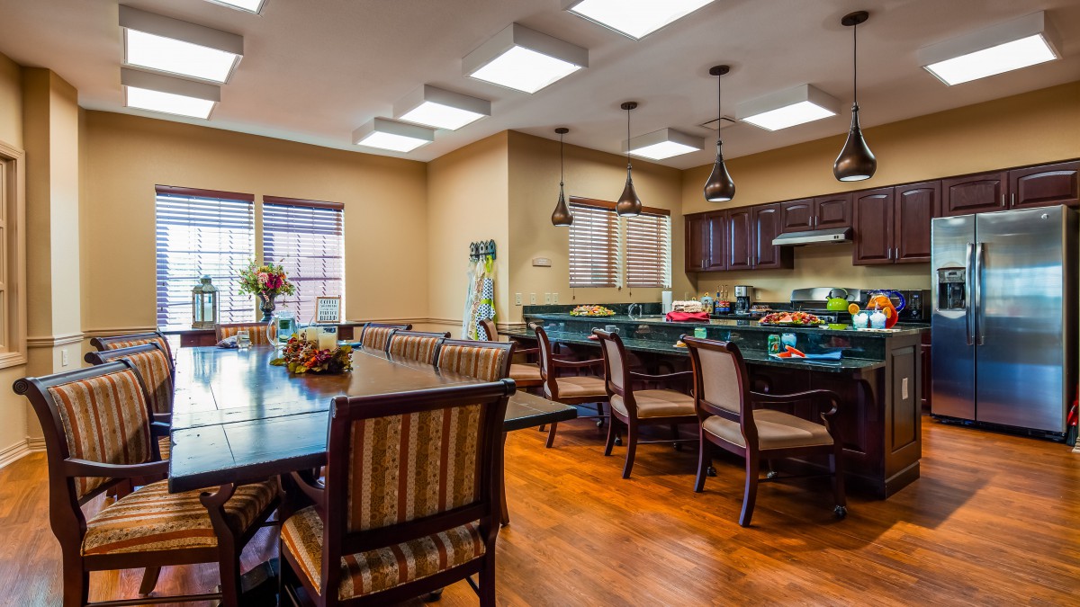 A cozy dining area with kitchen, featuring wooden tables and chairs, pendant lighting, and a hardwood floor.