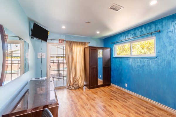 A bright room with blue walls, a wooden floor, and simple furnishings including a wardrobe, desk, and mounted TV.