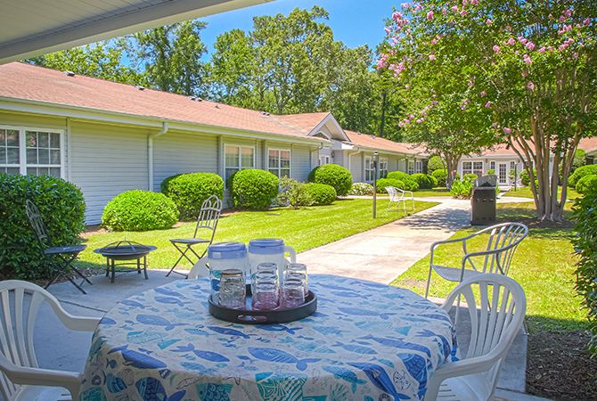 A sunny outdoor patio area with tables and chairs, surrounded by well-maintained lawns and buildings.