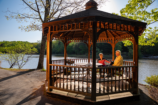 A serene afternoon in a gazebo by the lake, where two individuals enjoy a peaceful moment surrounded by nature.