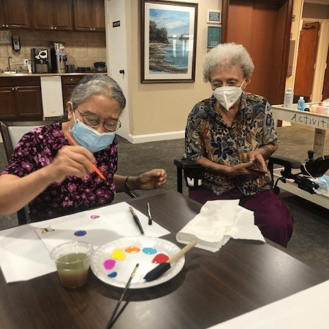 Two elderly women wearing masks engage in a painting activity indoors.