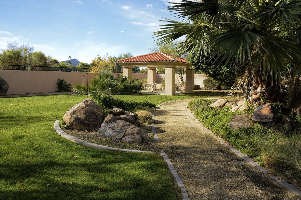 A serene garden featuring a paved pathway leading to a gazebo, surrounded by lush greenery and large rocks.