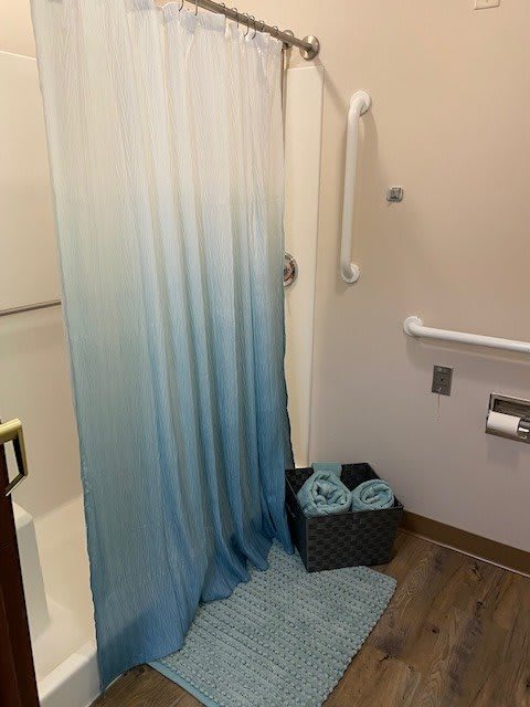 A bathroom with an ombre shower curtain, handrails, a basket of towels, and a matching rug.