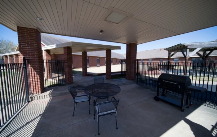 A covered patio with a table, chairs, and a grill next to a brick building.