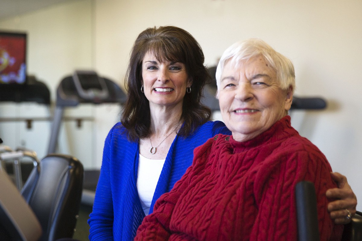 Two women smiling indoors, one wearing a blue cardigan and the other in a red sweater.