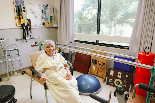 An elderly individual seated in a rehabilitation center, surrounded by various exercise equipment.