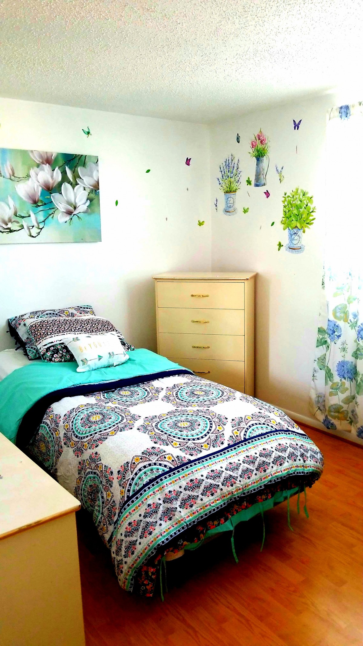 A cozy bedroom with a single bed, featuring colorful and floral bedding, complemented by wall art and a wooden dresser under natural light.
