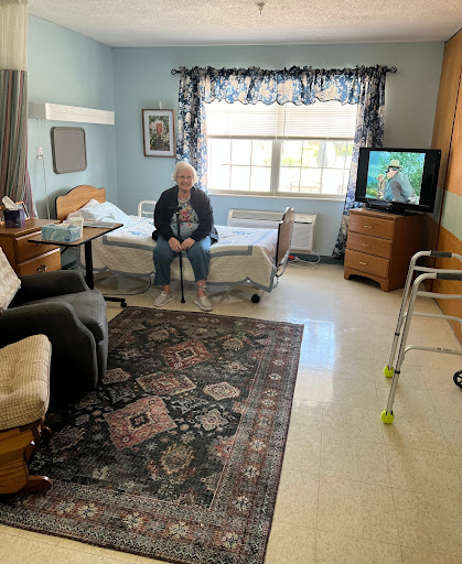 An elderly person sitting in a well-furnished hospital room with a view outside.