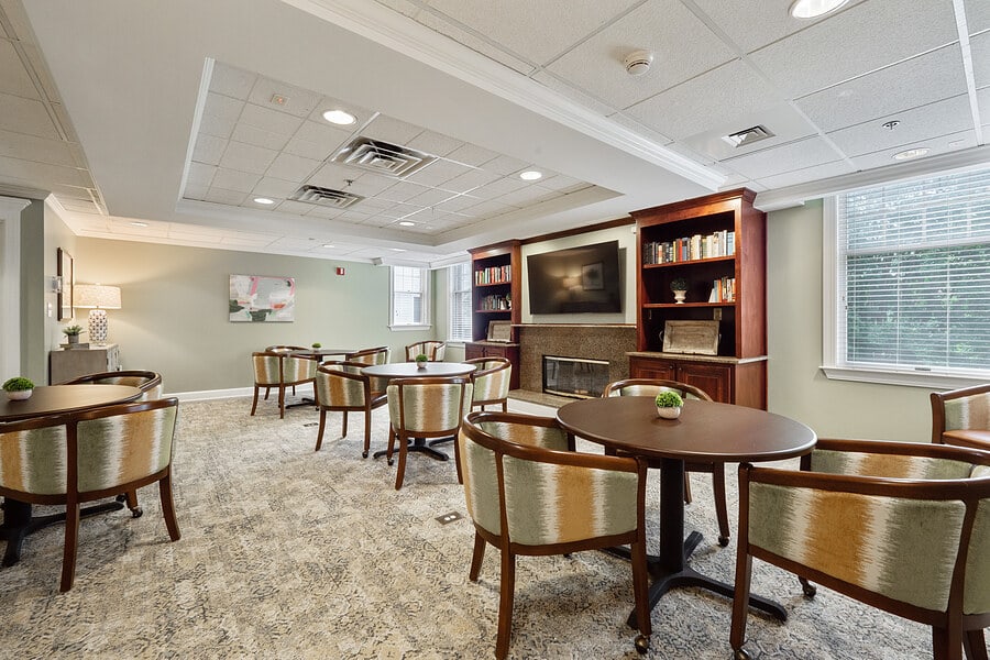 A cozy library room with tables and chairs, bookshelves, and a TV.
