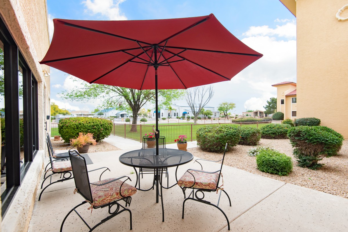 A patio with a red umbrella shading a table and chairs, surrounded by a landscaped garden on a sunny day.