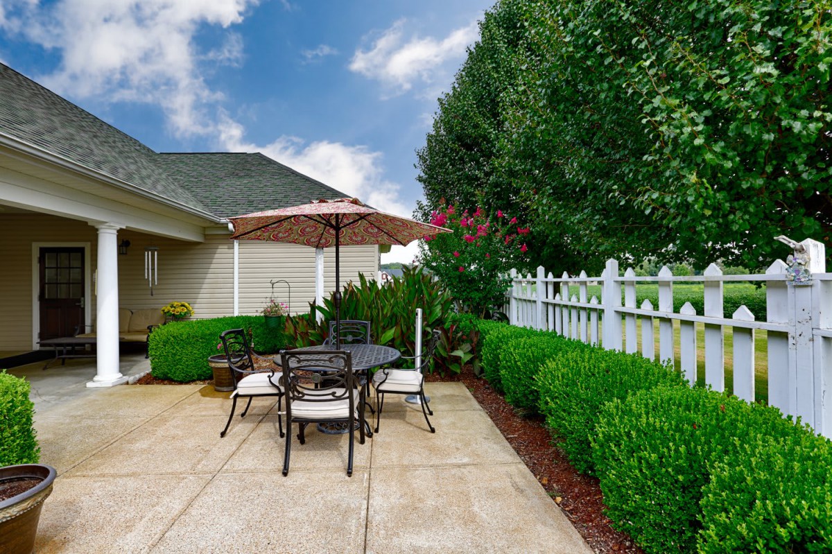 A cozy patio with outdoor seating, surrounded by lush greenery and a white fence.