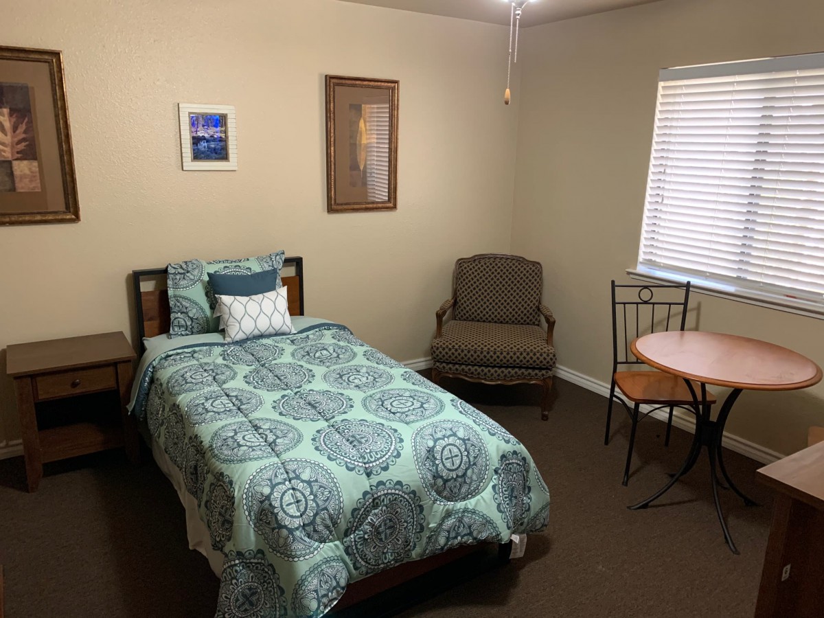 A cozy bedroom featuring a single bed with patterned bedding, an armchair, and a small table with a chair by the window.
