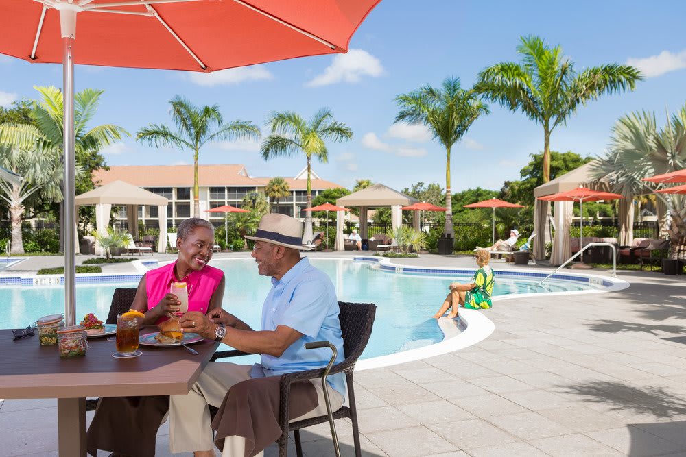 A senior couple enjoying a meal by the pool in a tropical resort setting.