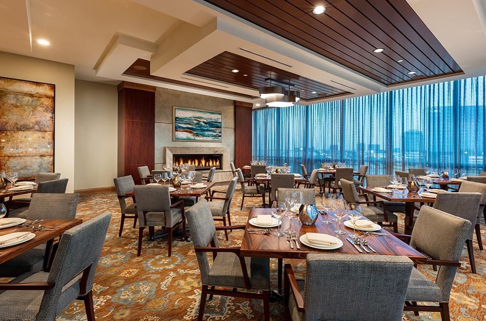 An elegant restaurant dining room with neatly arranged tables and chairs, featuring a fireplace and large windows.