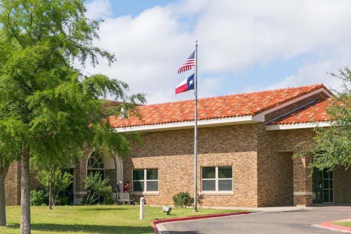 A brick building with American and Texas flags in front, surrounded by trees and under a cloudy sky.