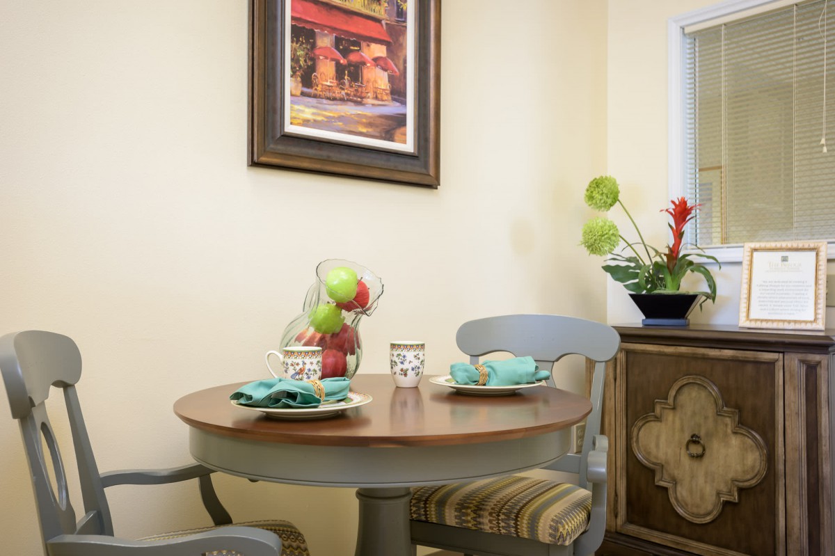A cozy dining nook with a round table set for two, featuring a decorative fruit bowl and framed artwork on the wall.