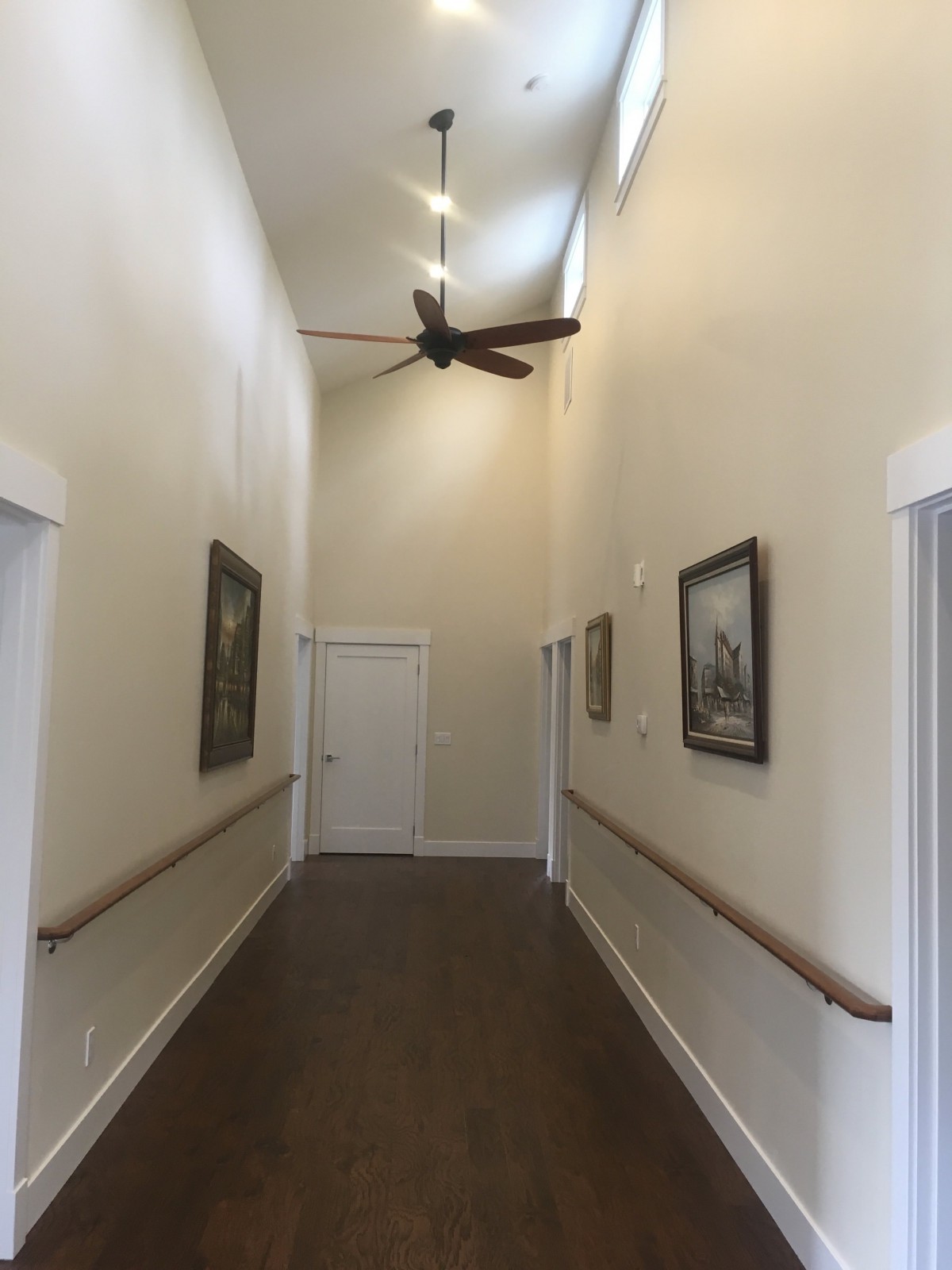 A long hallway with paintings, a ceiling fan, and handrails along the walls.