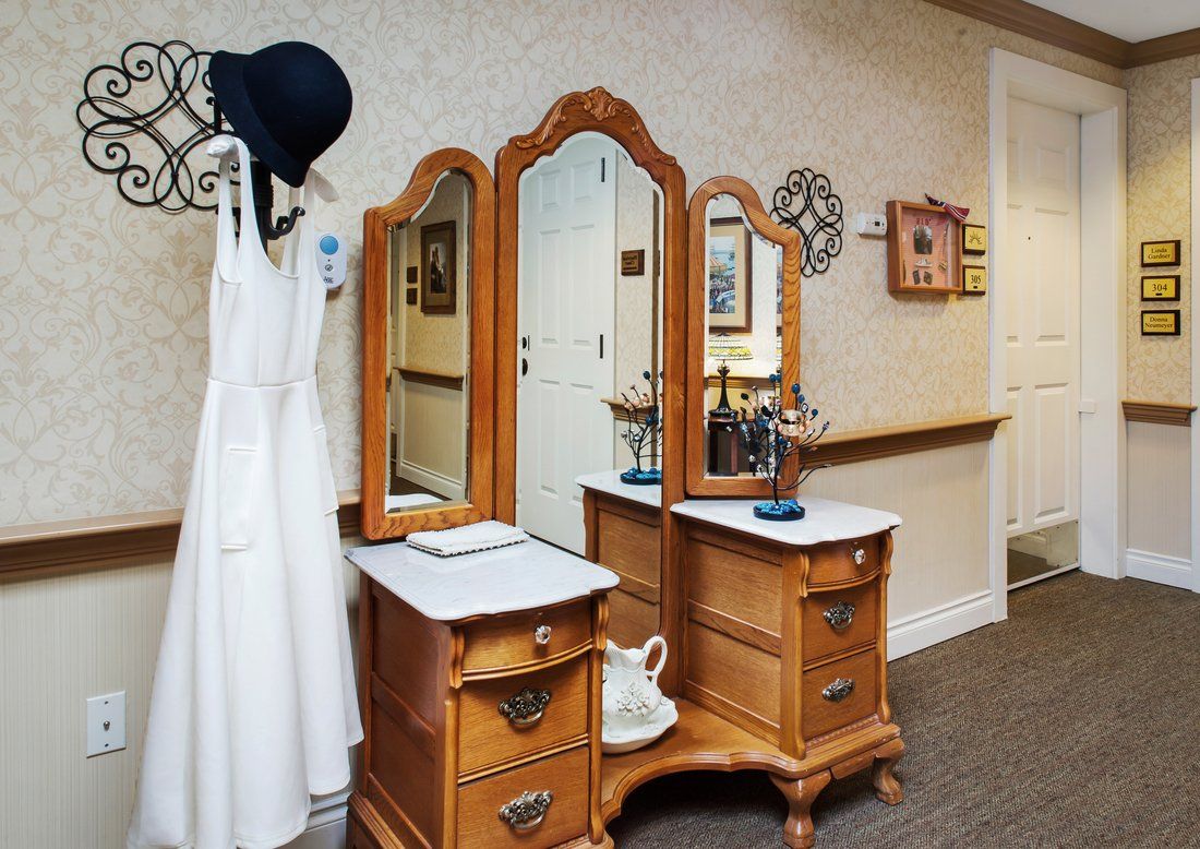 A vintage wooden dresser with a triple mirror, accompanied by a hanging hat and dress in an elegant interior setting.
