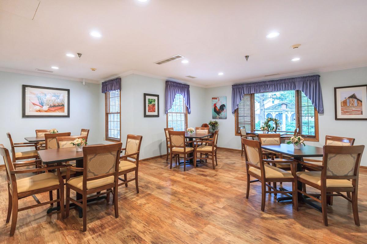 A well-lit dining room with tables and chairs, featuring hardwood floors and windows with curtains.