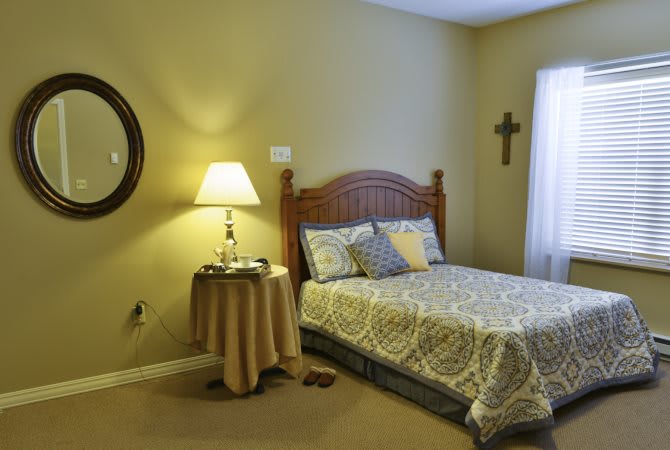 Cozy bedroom with warm lighting, featuring a bed with patterned linens and decorative pillows, a side table with a lamp, and wall decorations including a mirror and cross.