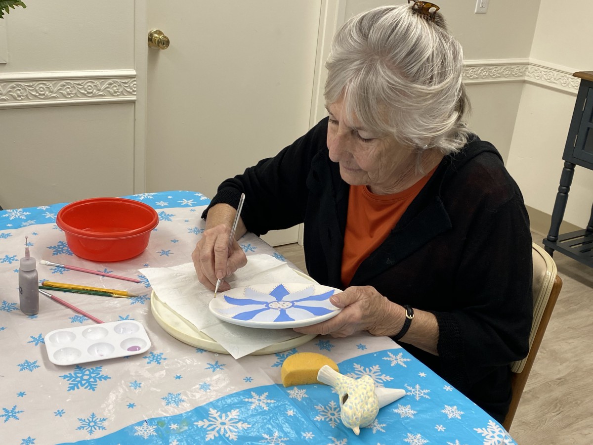 A woman is focused on painting a design on a plate at a table covered with a snowflake-patterned tablecloth.