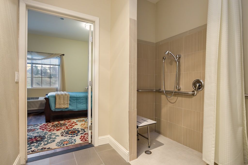 An accessible bathroom with a walk-in shower featuring grab bars and a shower chair, adjacent to a cozy bedroom with bright decor.