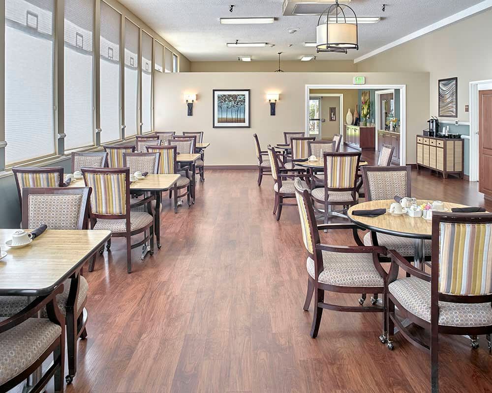 A bright and spacious dining area with neatly arranged tables and chairs, featuring wood flooring and large windows.