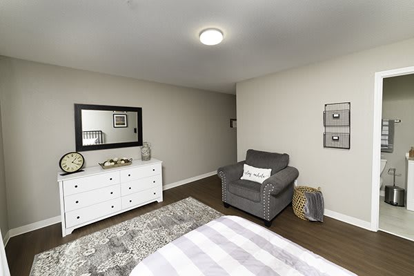 A cozy bedroom featuring a gray armchair, a white dresser with a mirror above, and decorative elements such as a rug and wall art.