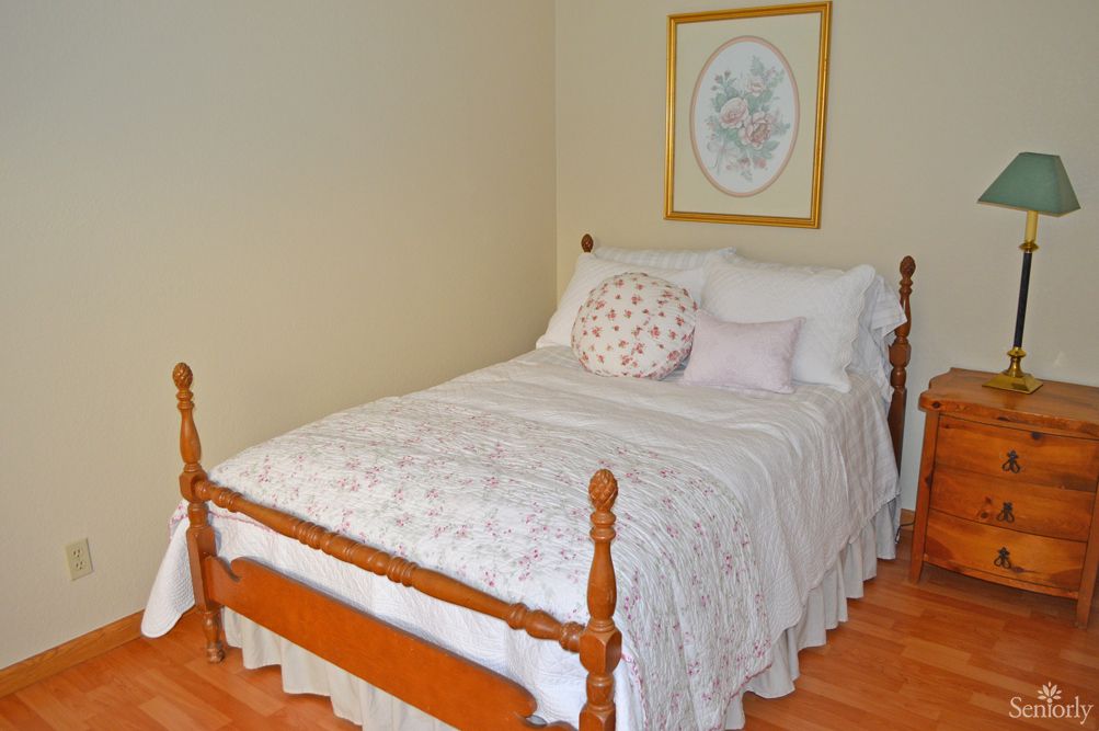 A cozy bedroom featuring a bed with a wooden frame, adorned with a quilt and pillows, and an artwork hanging on the wall.
