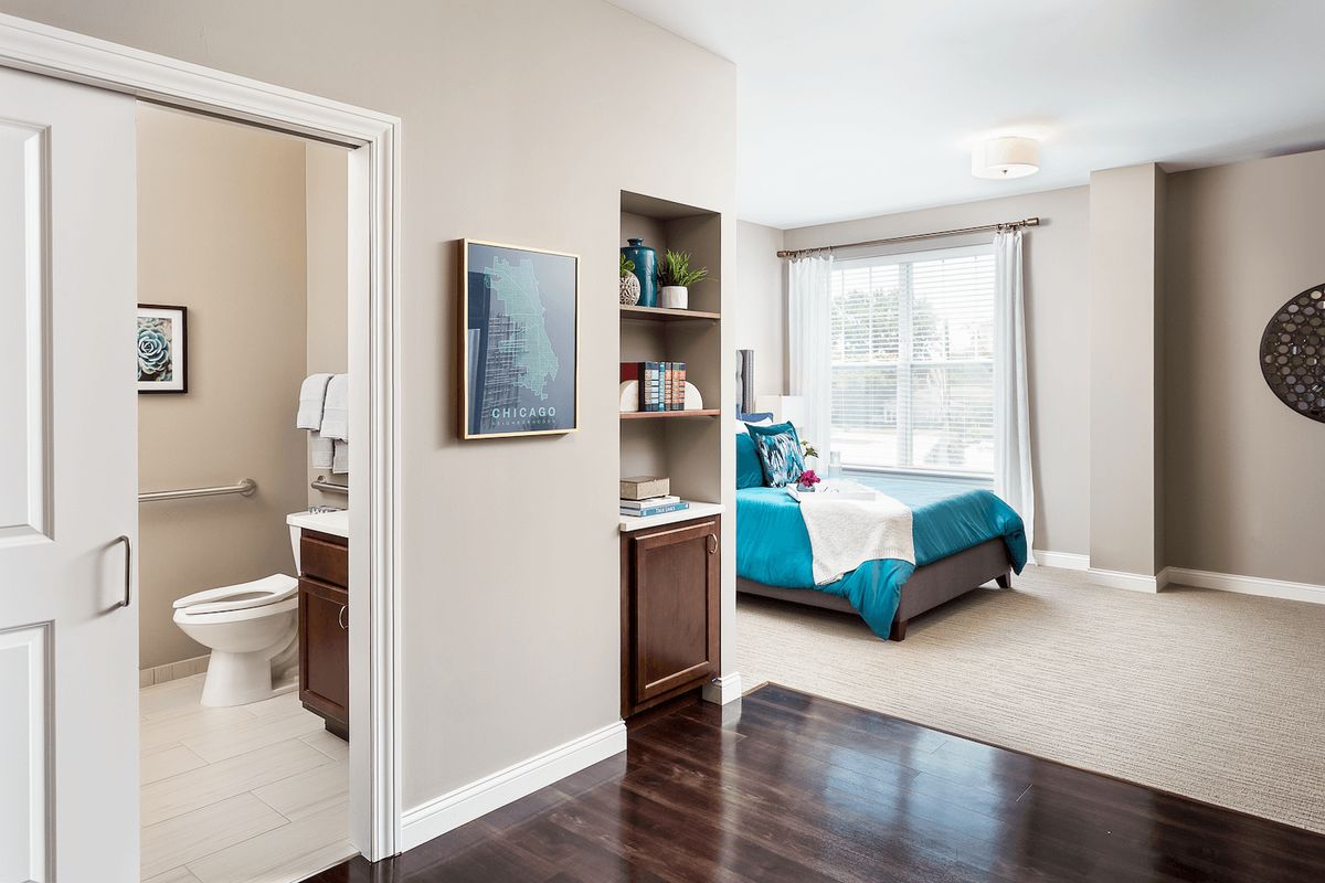 A modern bedroom with a view into an adjoining bathroom, featuring sleek furnishings and a stylish bookshelf.