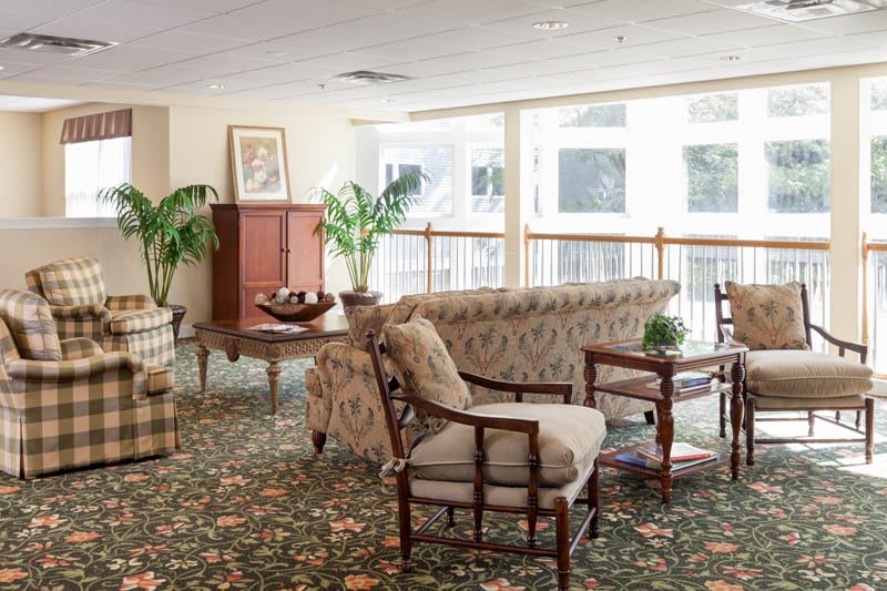 A bright and spacious living room with classic furniture, featuring a patterned sofa, chairs, wooden tables, and potted plants.