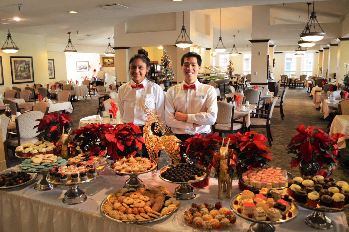 Two waiters stand behind a festive holiday dessert buffet in a decorated restaurant.