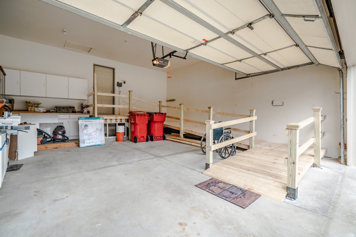 A spacious garage with a concrete floor, featuring a wheelchair ramp, storage shelves, and recycling bins.