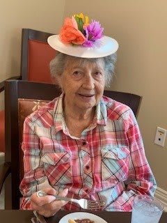An elderly woman wearing a plaid shirt and a colorful hat sits indoors on a chair.