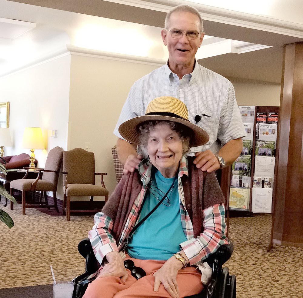 A smiling elderly woman in a wheelchair, wearing a hat, with a man standing behind her in an indoor setting.
