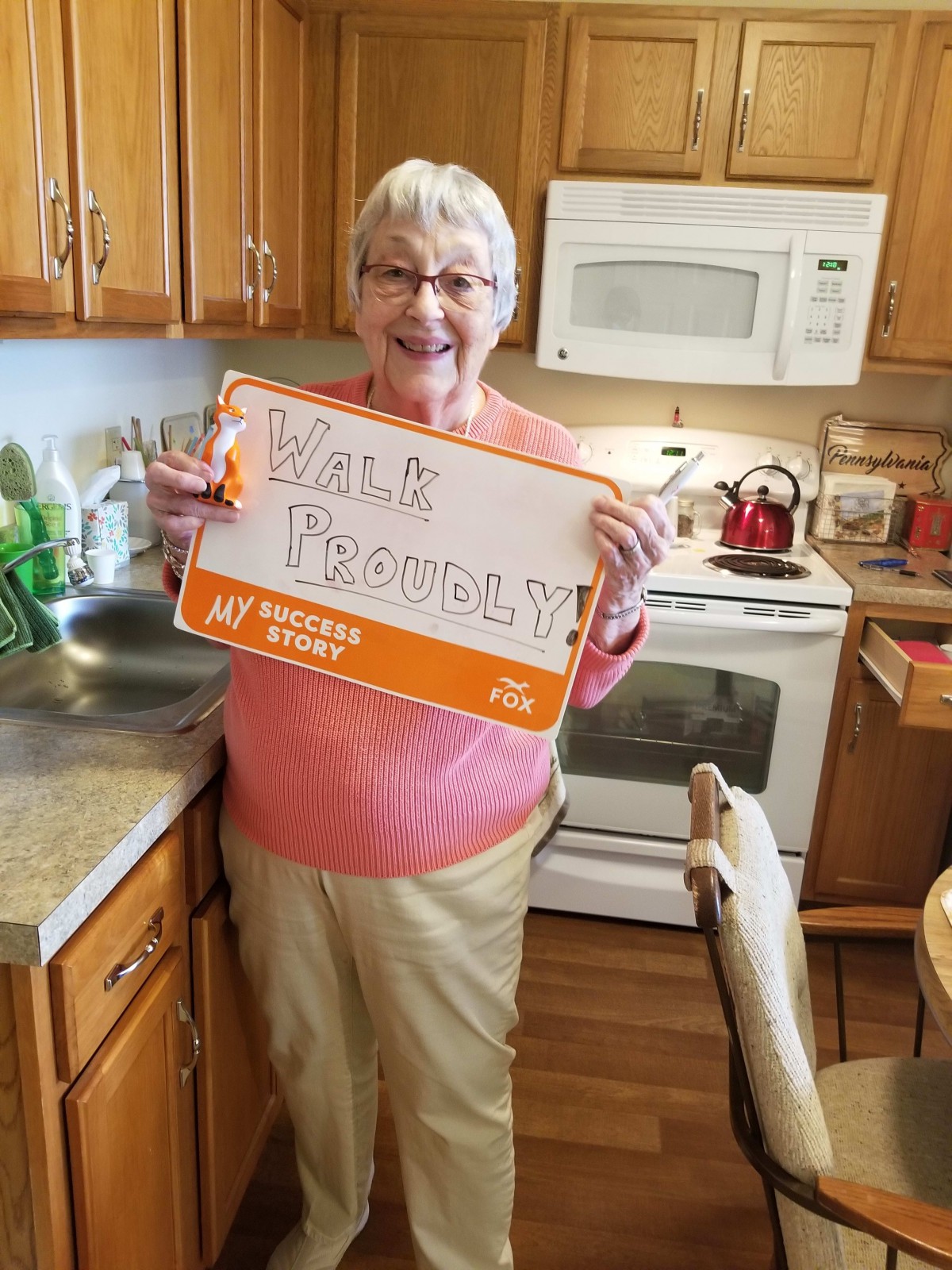 An elderly woman stands in her kitchen holding a sign with 'Birth Freedom' written on it.