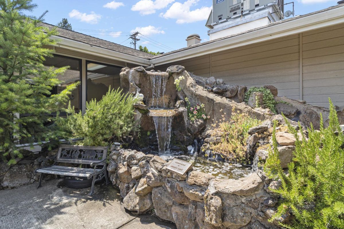 A small outdoor garden featuring a waterfall fountain surrounded by rocks and lush greenery, with a bench nearby.