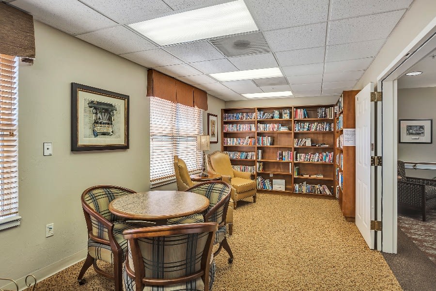 A cozy library room with bookshelves, a reading area featuring an armchair, and a table with chairs.