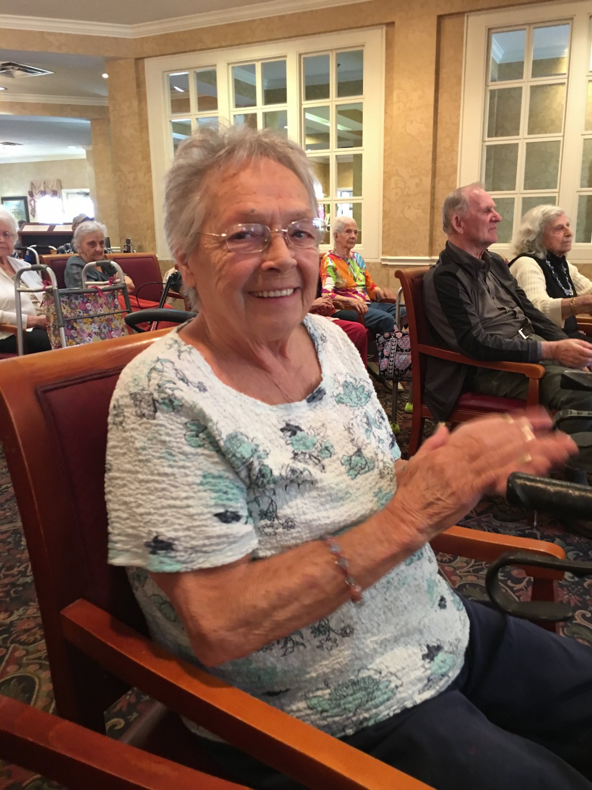 An elderly woman clapping while seated indoors, smiling as she enjoys an event.