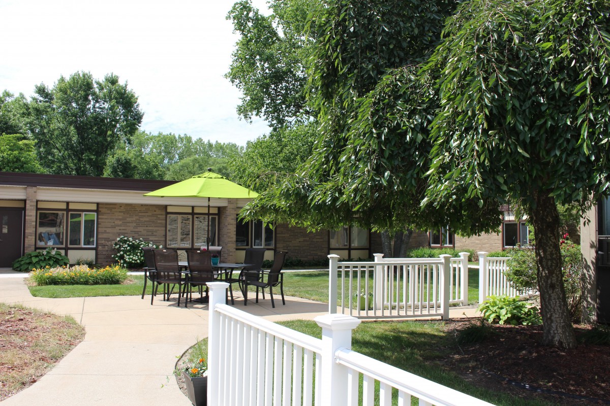 A serene outdoor patio area with a table, chairs, and a green umbrella, surrounded by trees and a white fence.