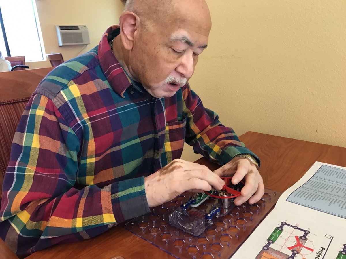 An elderly man wearing a plaid shirt is focused on solving a puzzle at the table.