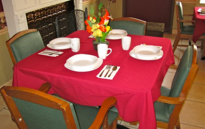 A neatly set dining table with a red tablecloth, featuring plates, mugs, cutlery, and a floral centerpiece.