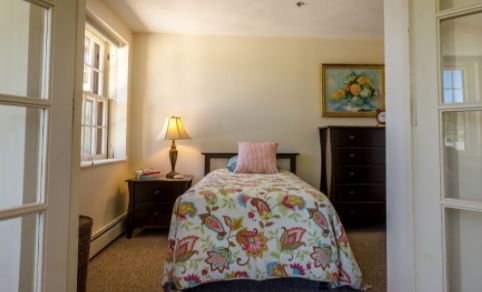 A cozy bedroom with a single bed featuring floral bedding, flanked by a lamp on a nightstand and a dresser with artwork above.