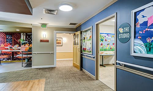 A well-lit interior hallway of a senior living facility with decorated walls and carpeted flooring.