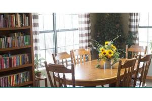 A cozy dining area with a wooden table and chairs, adorned with a vase of sunflowers. Natural light streams in through the window, and a bookshelf lines the wall.