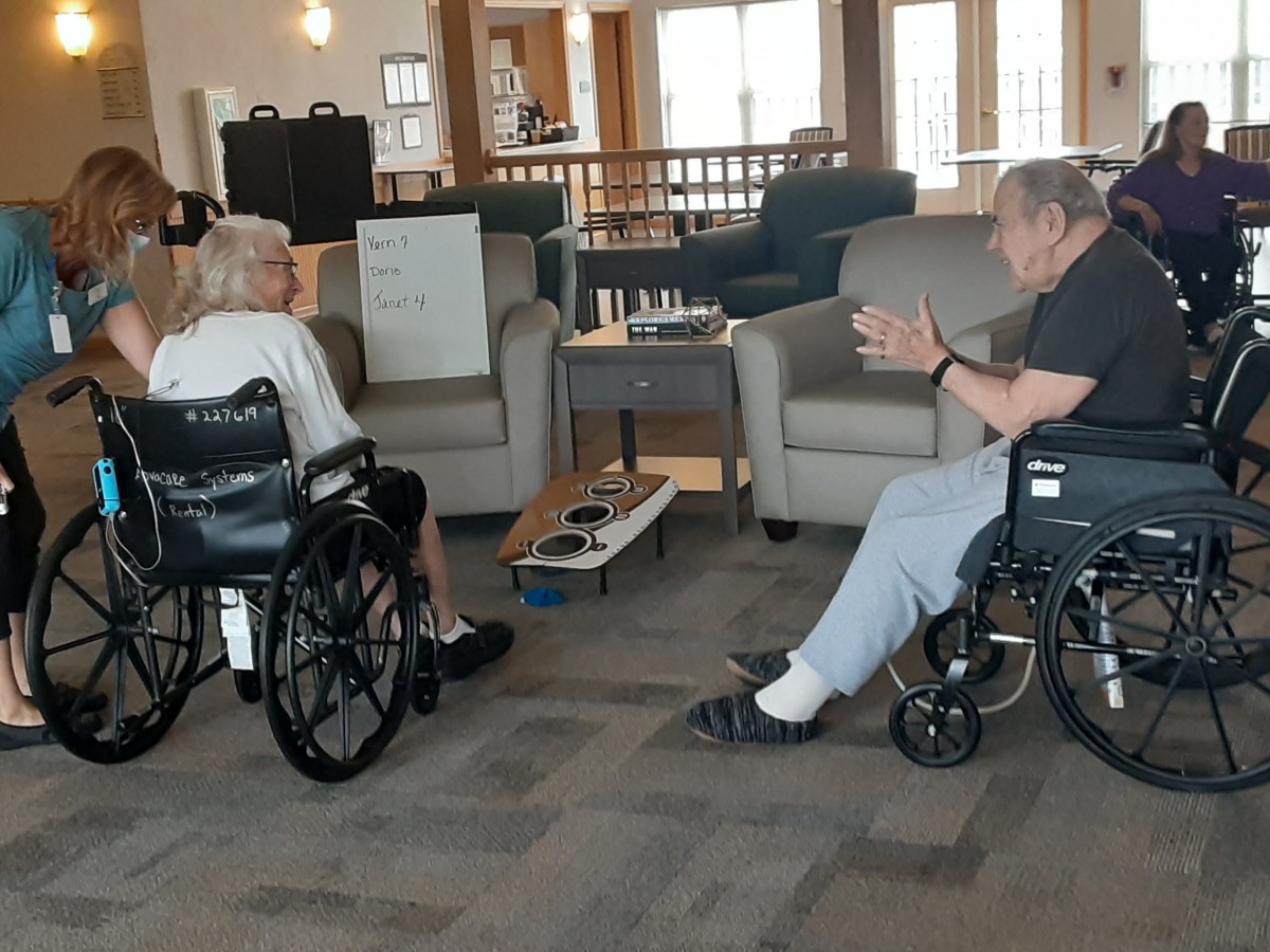 Two elderly individuals in wheelchairs engage in conversation inside a care facility living room.