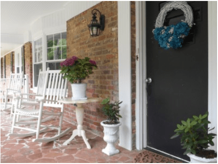 A cozy porch featuring a white rocking chair, potted plants, and a decorative wreath on the front door.