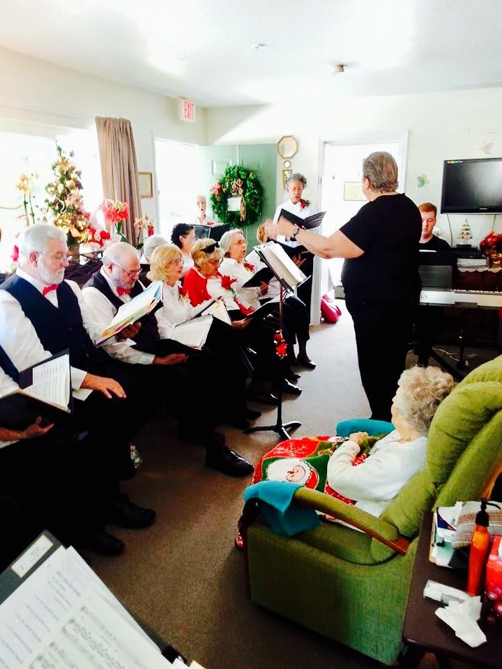 A choir of elderly individuals performs under the direction of a conductor, surrounded by festive holiday decorations.