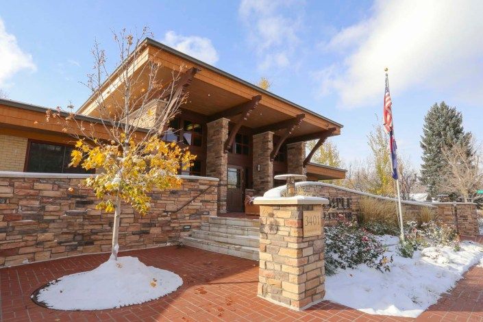 A modern brick building surrounded by snow, featuring an American flag and trees with autumn leaves.